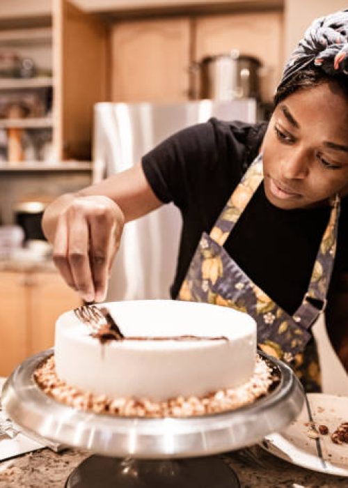 Young black woman baking a sweet cake from the scratch in her kitchen. She is putting icing on the new cake.  Interior of private home.